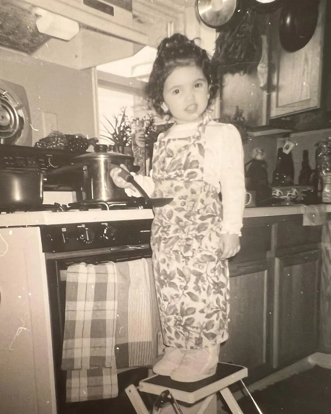 Ari as a little girl in an apron, standing at the stove
